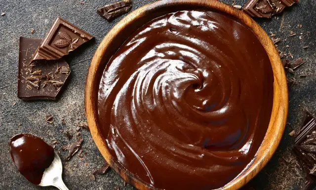Un bol en bois déborde de chocolat fondu, prêt à faire du chocolat. Des éclats de chocolat noir et une cuillère en argent complètent la scène gourmande.