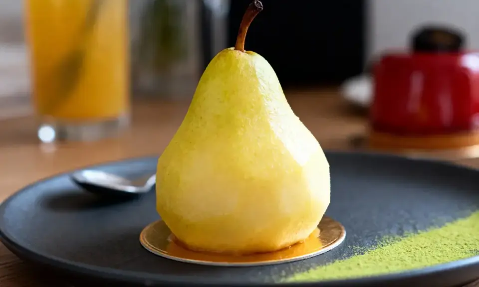 Un entremet poire, glacé et brillant, repose sur une assiette noire avec une touche de poudre verte.