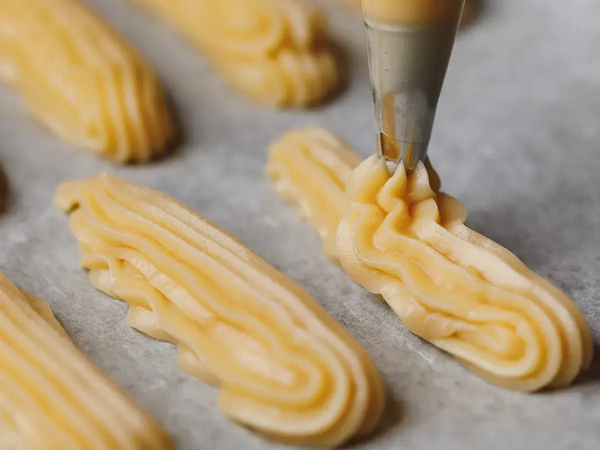 Préparation de la pâte à choux pour la recette de maman choux à la crème. La poche à douille dépose des éclairs sur du papier cuisson.