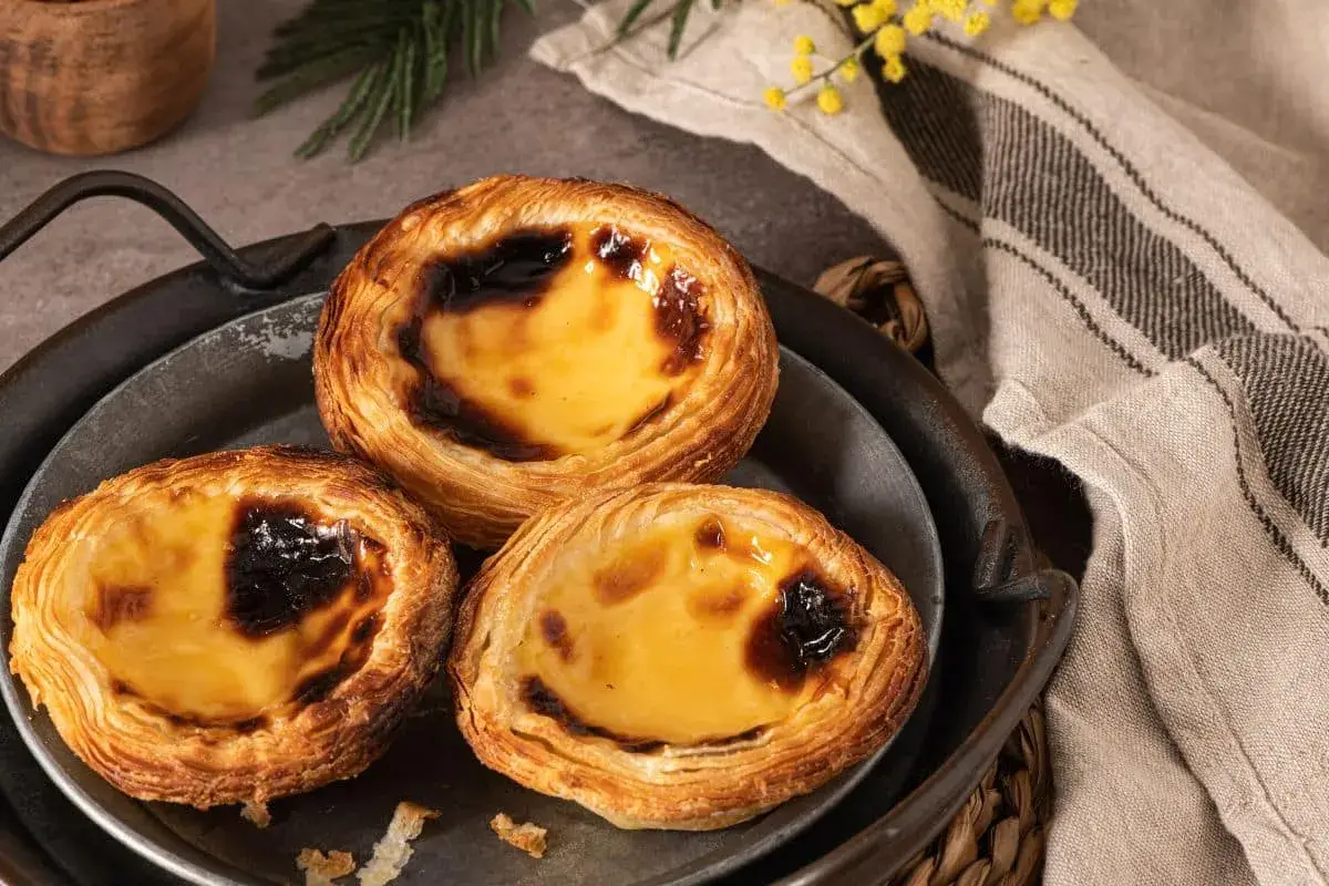 Trois délicieux pasteis de nata dorés, leur crème onctueuse légèrement caramélisée, sur un plateau en métal.