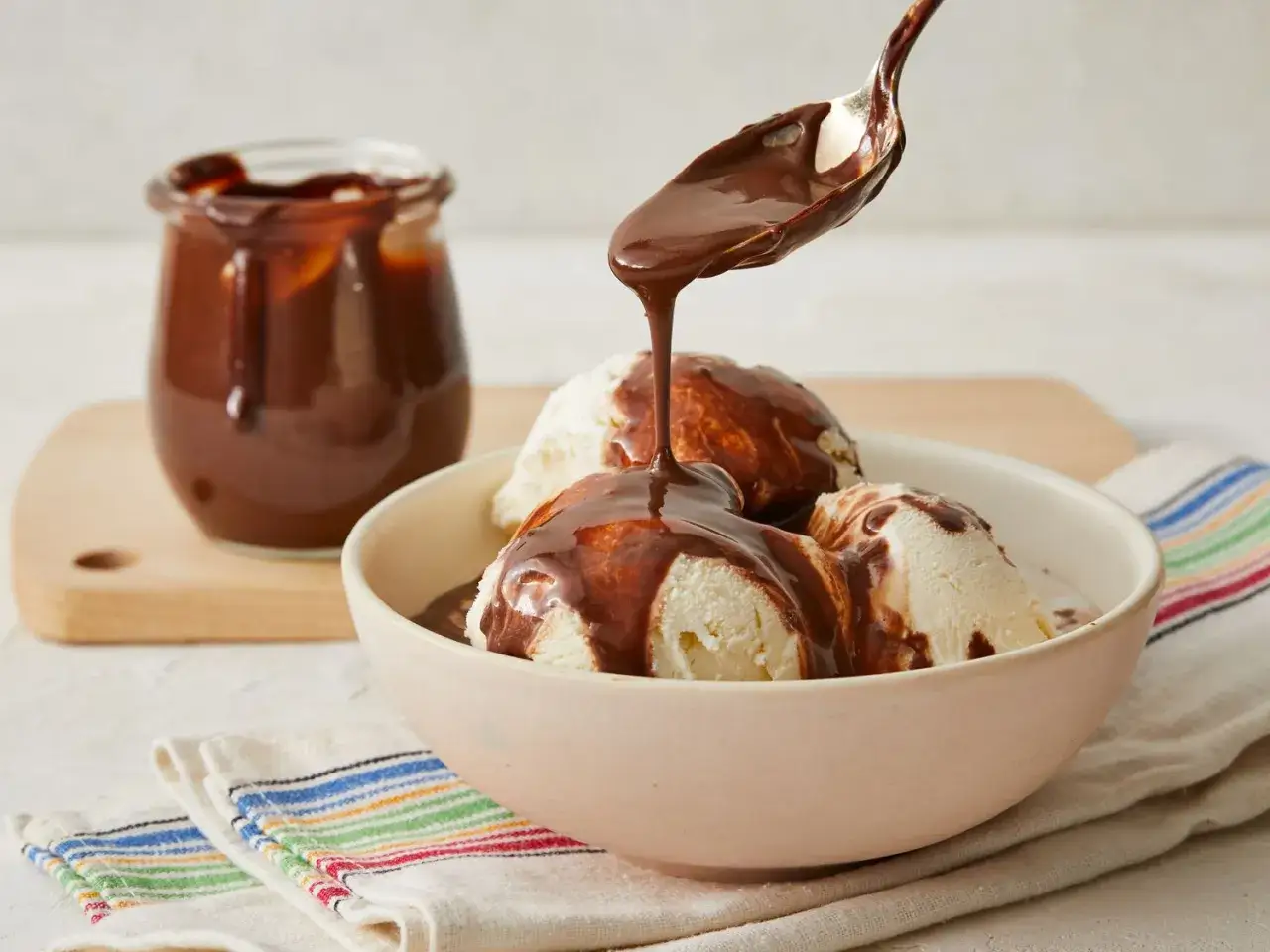 Une cuill&egrave;re verse une sauce chocolat qui ne fige pas sur des boules de glace vanille dans un bol. Un pot de sauce est visible en arri&egrave;re-plan.