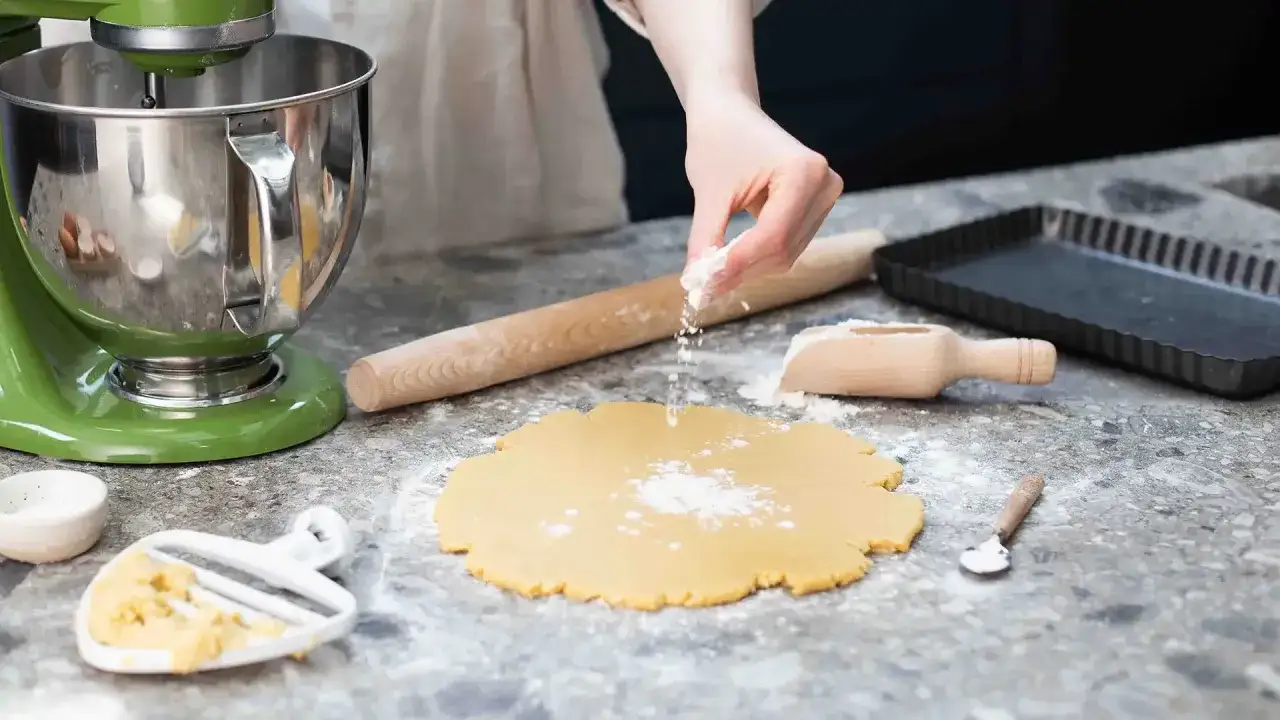 Préparation d'une pâte sablée au robot. Une main saupoudre de la farine sur la pâte étalée, à côté d'un rouleau à pâtisserie et d'un moule à tarte.