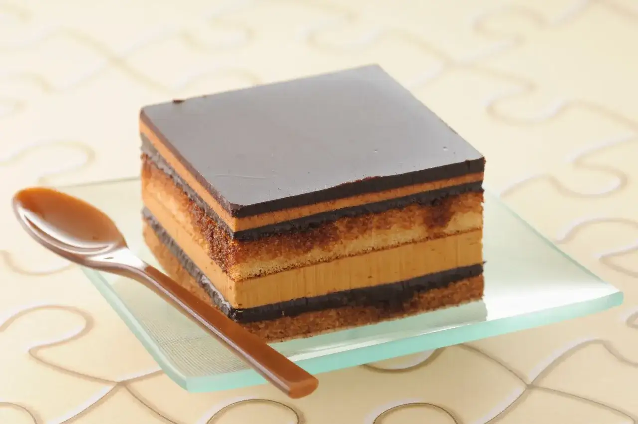 Un carré de gâteau opéra aux couches de biscuit, ganache et crème au café, sur une assiette en verre avec une cuillère.