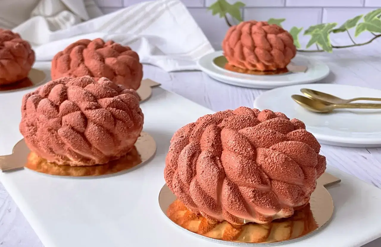 Plusieurs dacquoises noisette aux formes originales, recouvertes d'une poudre orangée, sont présentées sur des socles dorés.