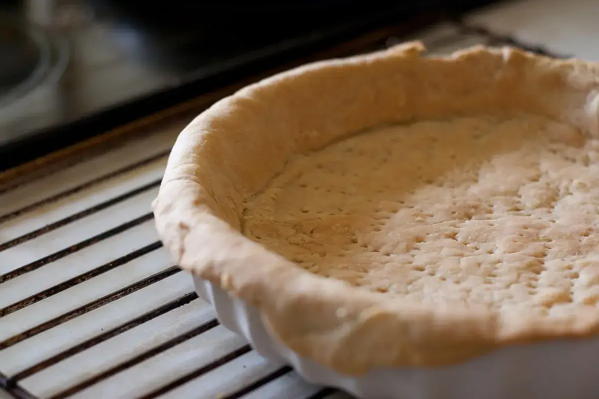 Pâte brisée prête pour la cuisson à blanc, posée sur une grille. Elle est légèrement dorée et percée de petits trous.
