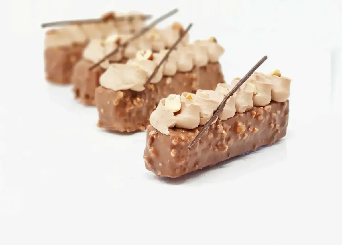 Petits gâteaux rectangulaires enrobés de chocolat et de noisettes concassées, surmontés d'une crème légère et d'un bâtonnet de chocolat. Le croustillant praliné promet une texture divine.