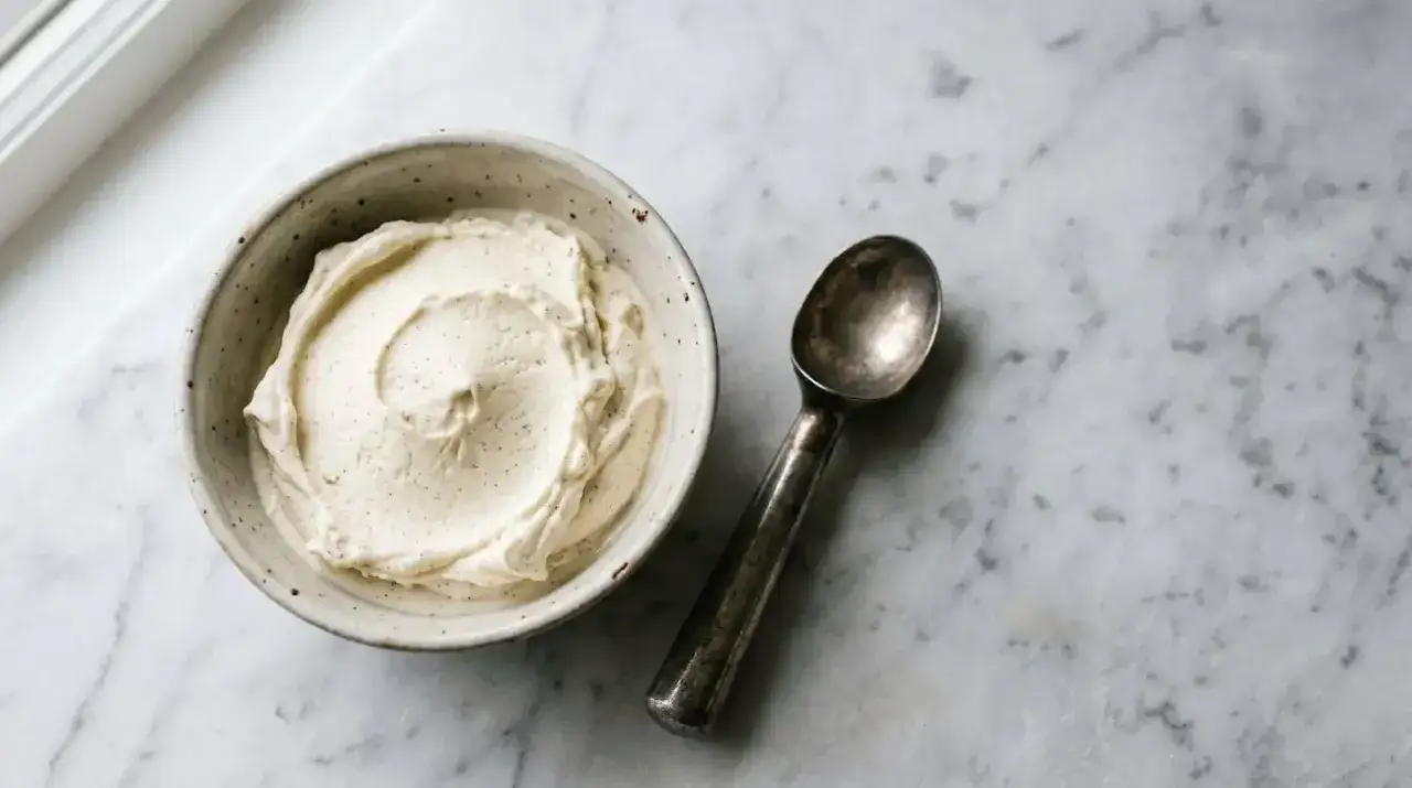 Un bol de glace vanille maison, parsemé de points de vanille, à côté d'une cuillère à glace sur un marbre.