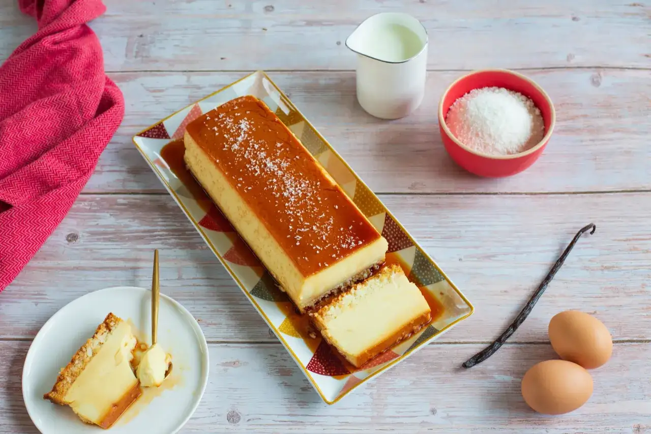 Un délicieux flan coco, saupoudré de noix de coco râpée, est prêt à être dégusté. Des ingrédients frais comme des œufs et une gousse de vanille sont à proximité.