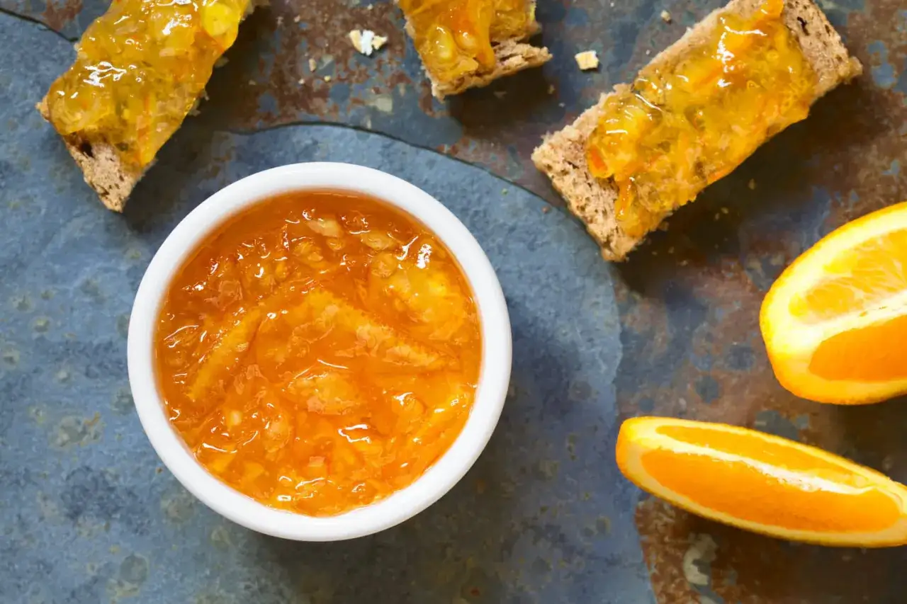 Confiture d'écorce d'orange brillante dans un bol blanc, servie sur des toasts avec des quartiers d'orange frais.