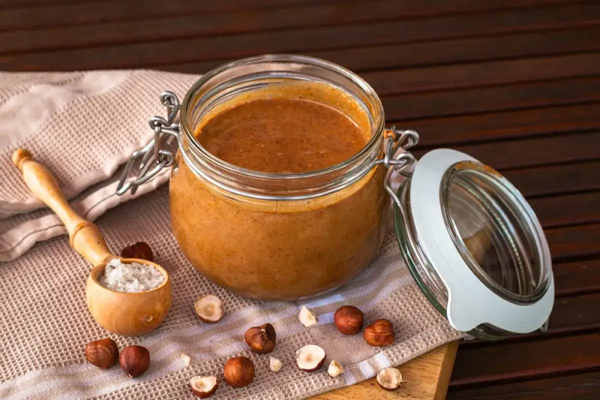 Un pot de praliné maison onctueux, entouré de noisettes entières et brisées, avec une cuillère en bois remplie de gros sel.