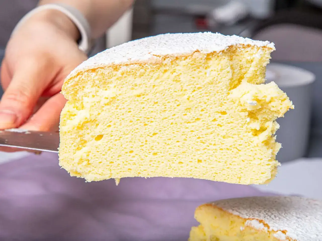 Une main coupe une tranche aérienne de cheesecake japonais saupoudré de sucre glace. Sa texture légère et moelleuse invite à la dégustation.