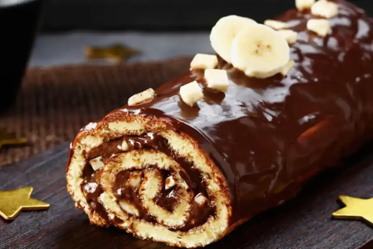 B&ucirc;che roul&eacute;e g&eacute;n&eacute;reusement napp&eacute;e d'un glacage chocolat caramel, garnie de rondelles de banane et de morceaux de noisettes.