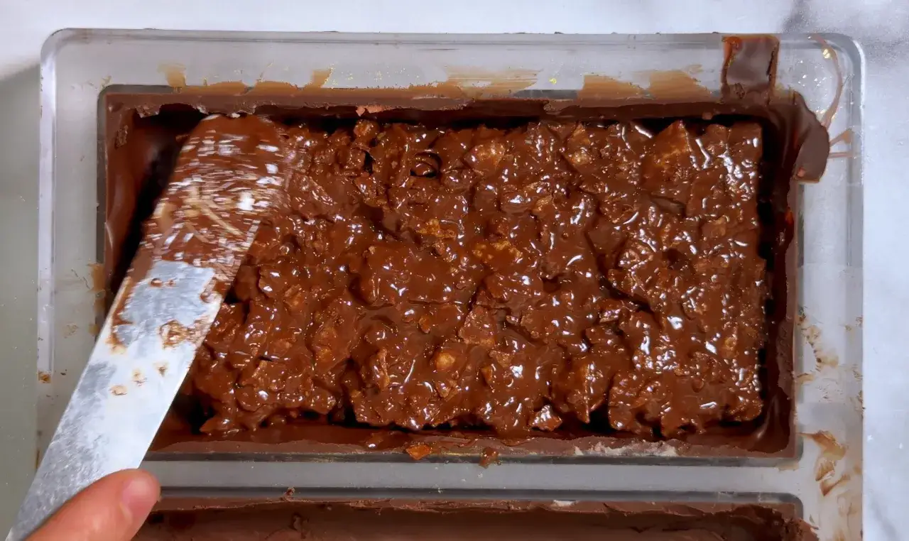 Une spatule lisse le dessus d'un mélange de chocolat et de céréales, créant une texture croustillant chocolat irrésistible dans un moule.