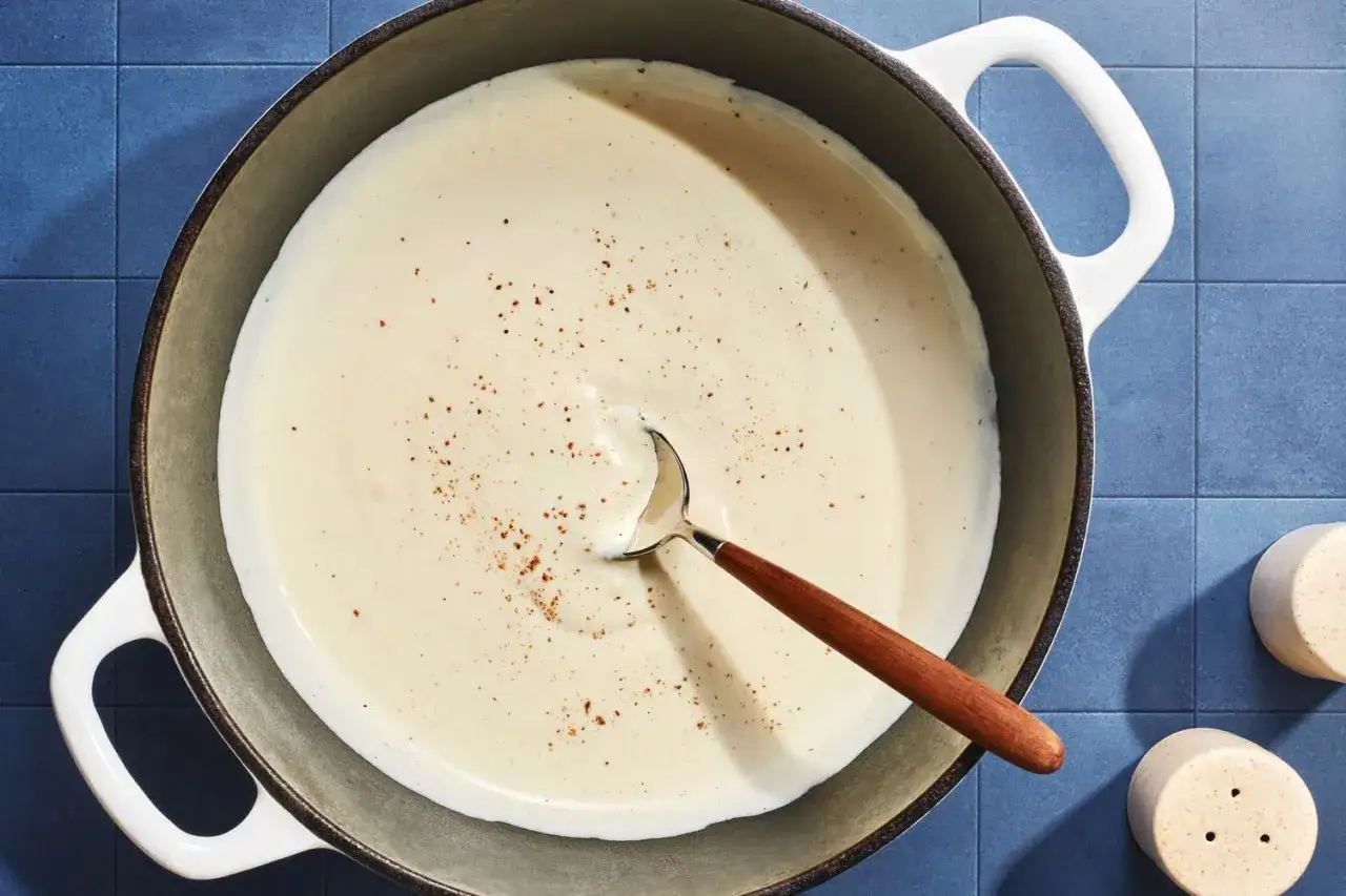 Une cuill&egrave;re en bois repose dans une casserole remplie d'une sauce cr&eacute;meuse, saupoudr&eacute;e de poivre noir.