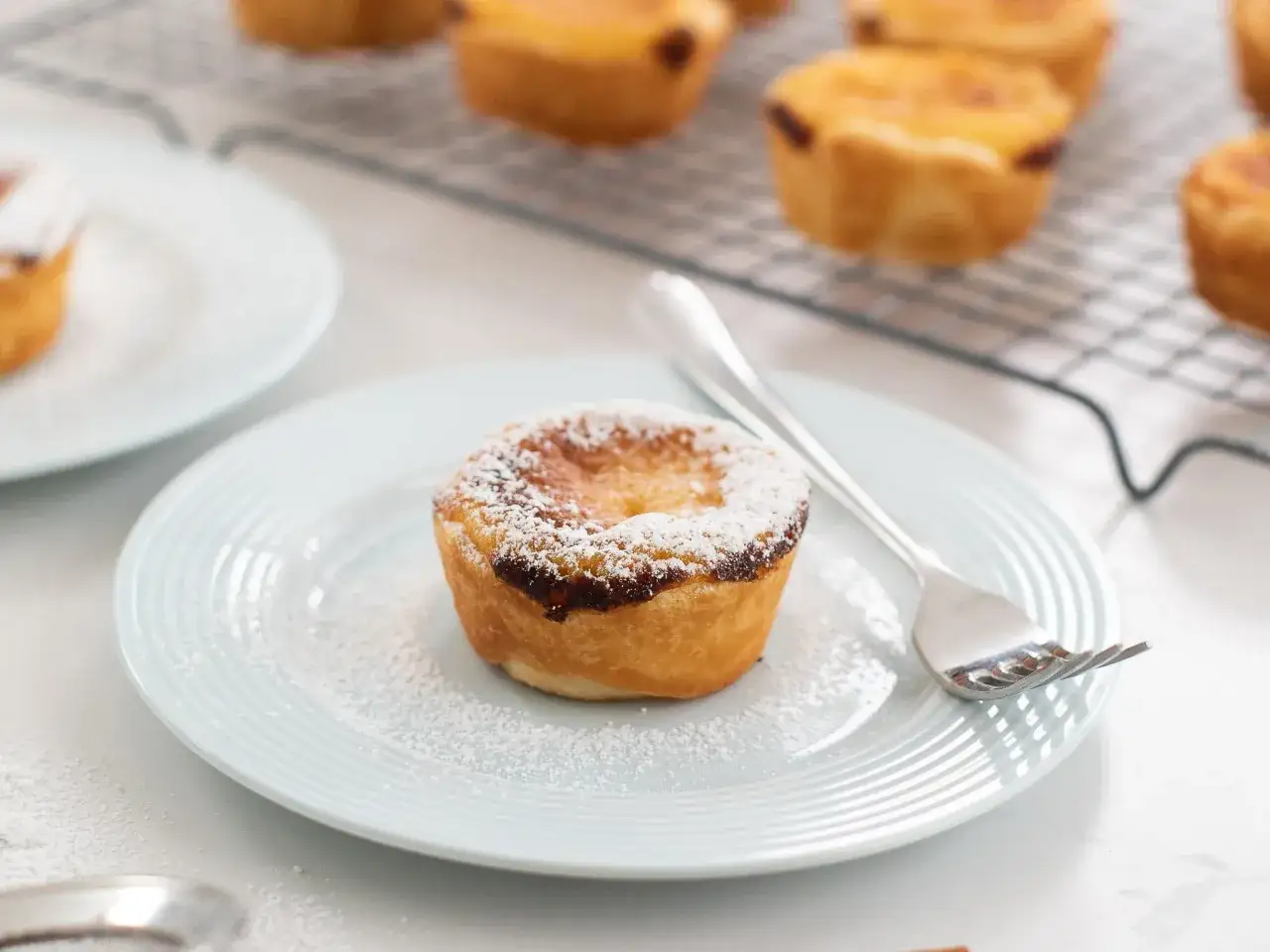 Un délicieux pasteis de nata, saupoudré de sucre glace, sur une assiette bleue avec une fourchette. D'autres sont sur une grille derrière.