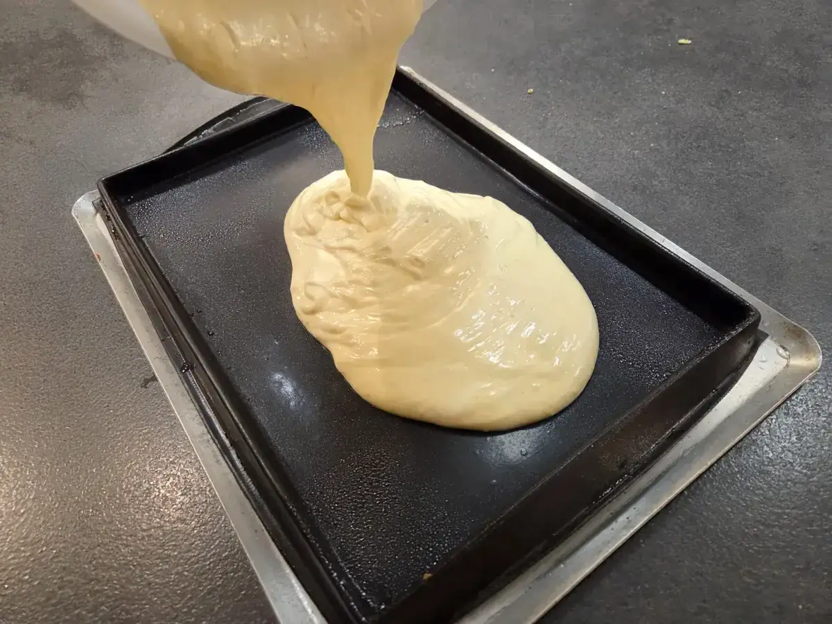 Une pâte onctueuse est versée dans un moule carré, formant le fond biscuit pour gâteau.