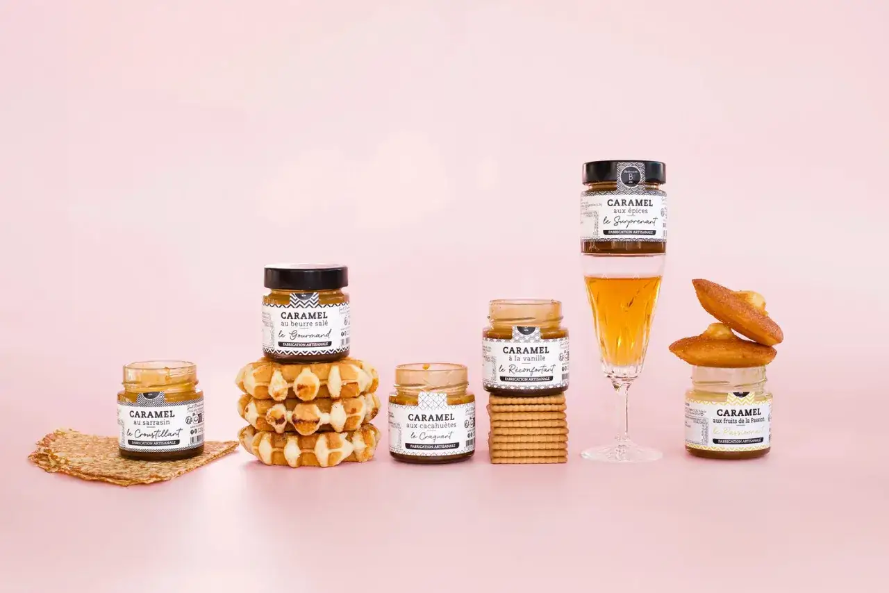Des pots de caramel artisanal, dont un caramel au beurre salé, côtoient gaufres, biscuits et madeleines sur fond rose.