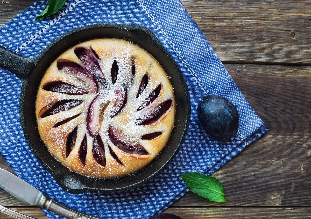 Clafoutis aux pruneaux, saupoudré de sucre glace, dans une poêle en fonte sur une serviette bleue.