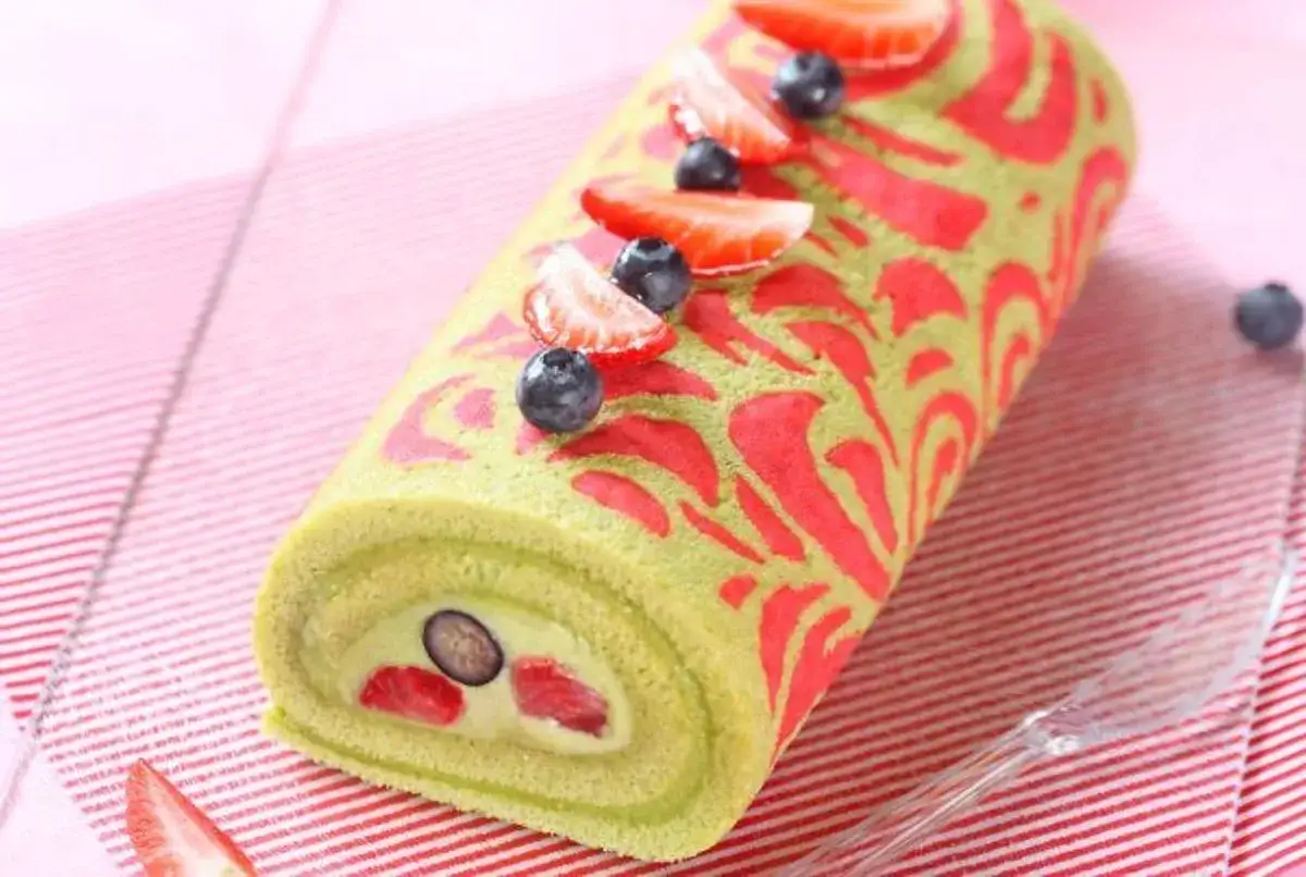 Bûche roulée verte décorée de motifs rouges, garnie de crème, fraises et myrtilles. Une belle idée pour une recette buche pistache framboise.