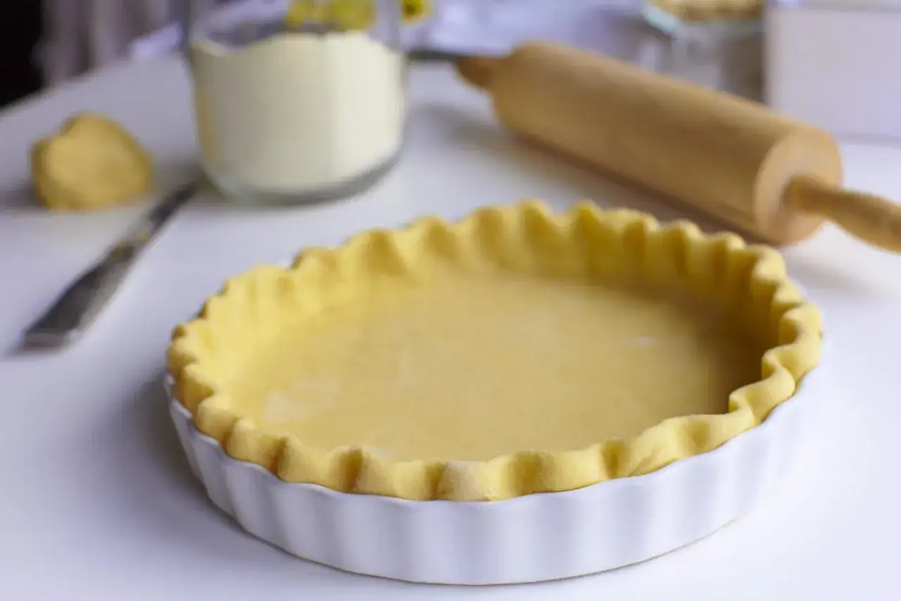 Pâte brisée prête à être garnie, un classique pour une recette facile. Rouleau à pâtisserie et ingrédients en arrière-plan.