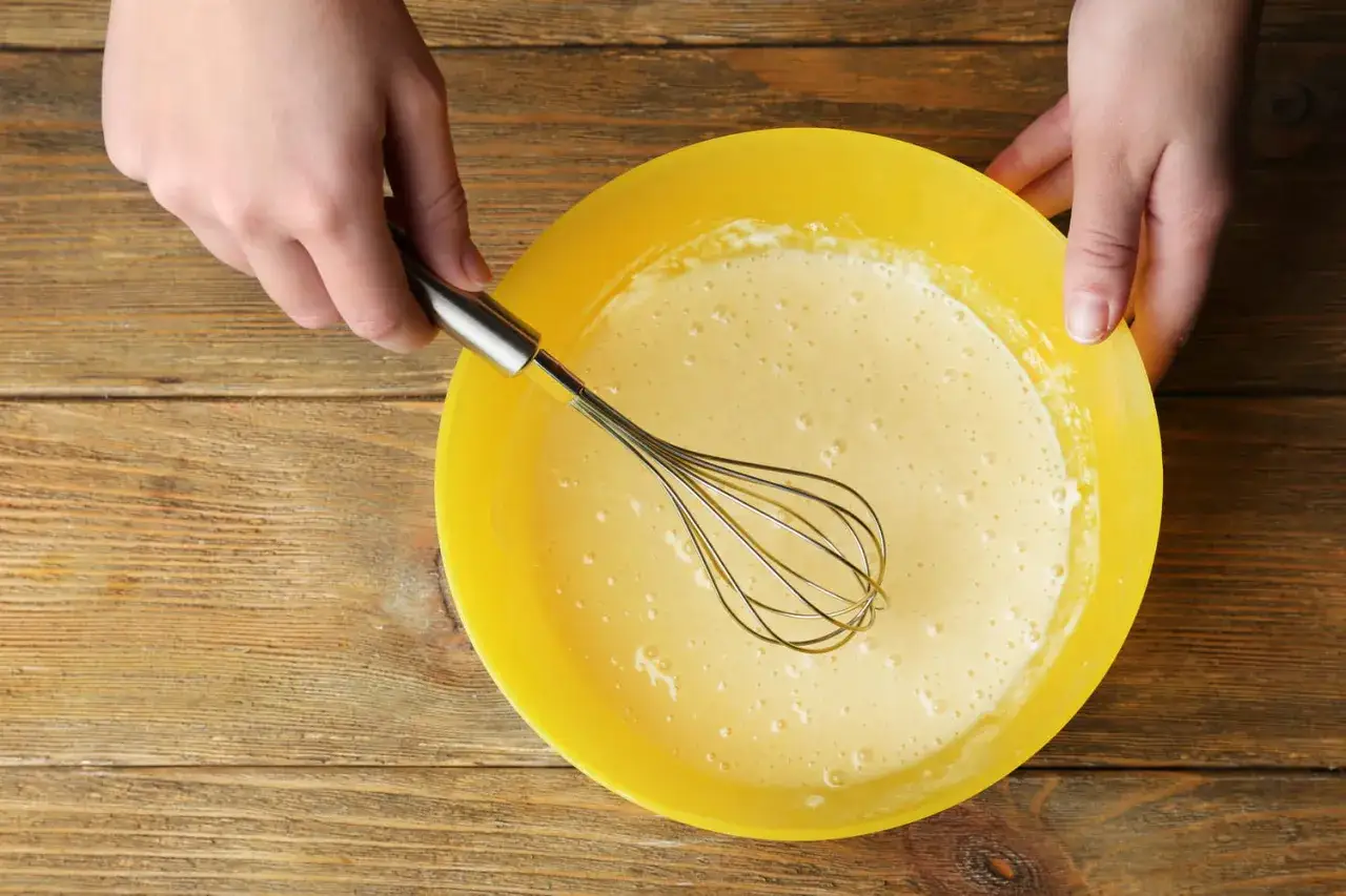 Mains préparant la pâte à crêpes dans un bol jaune. La pâte est prête pour son temps de repos.
