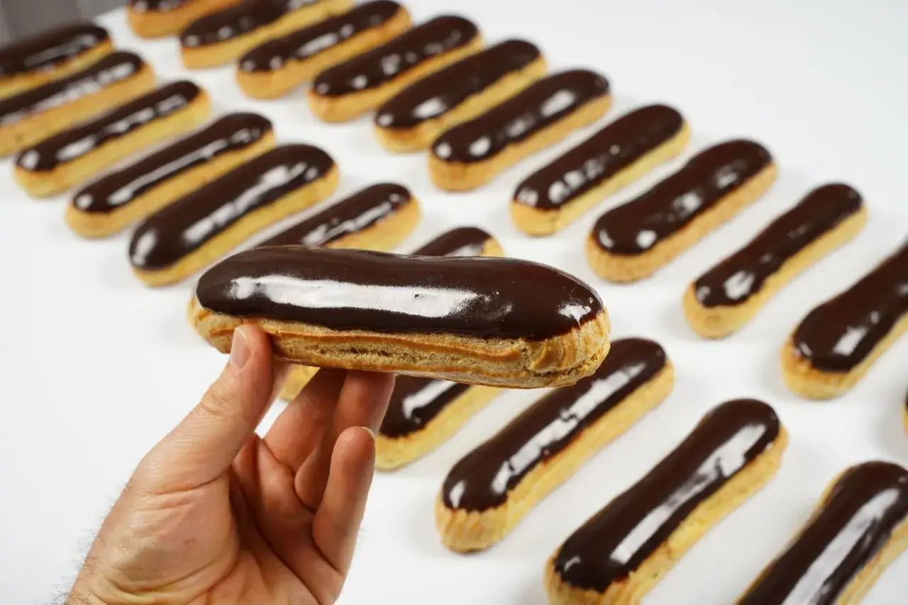 Une main tient un &eacute;clair au chocolat, avec une rang&eacute;e d'autres &eacute;clairs au chocolat brillants en arri&egrave;re-plan.