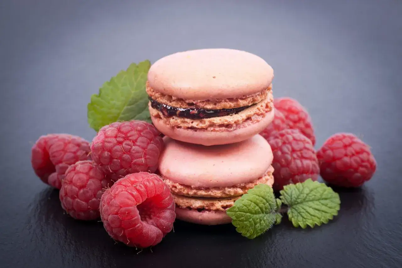 Deux macarons roses empilés, entourés de framboises fraîches et de feuilles de menthe, évoquent la douceur d'un macaron framboise.