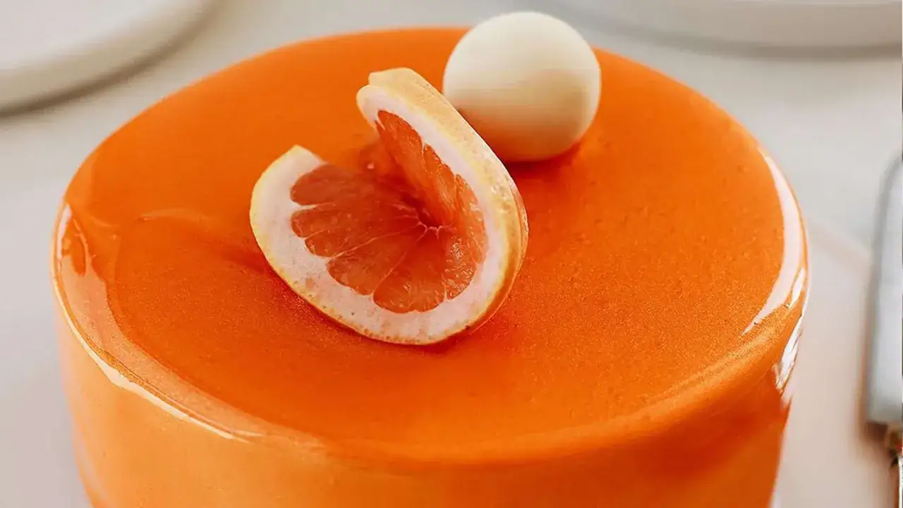 Gâteau rond au glaçage miroir orange vif, décoré d'une tranche de pamplemousse et d'une sphère blanche.