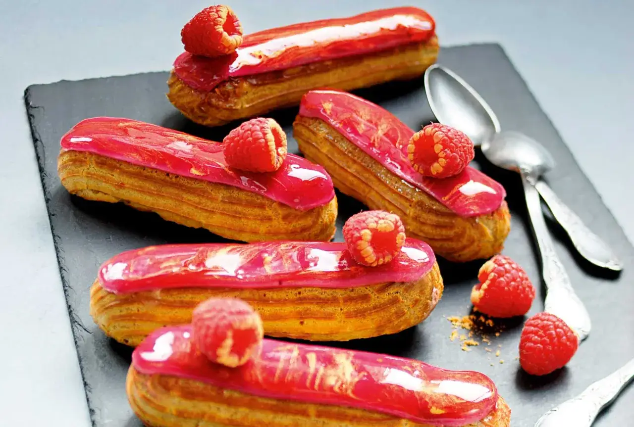 Cinq éclairs à la framboise, glacés de rose vif et ornés de fruits rouges frais, sur une ardoise.