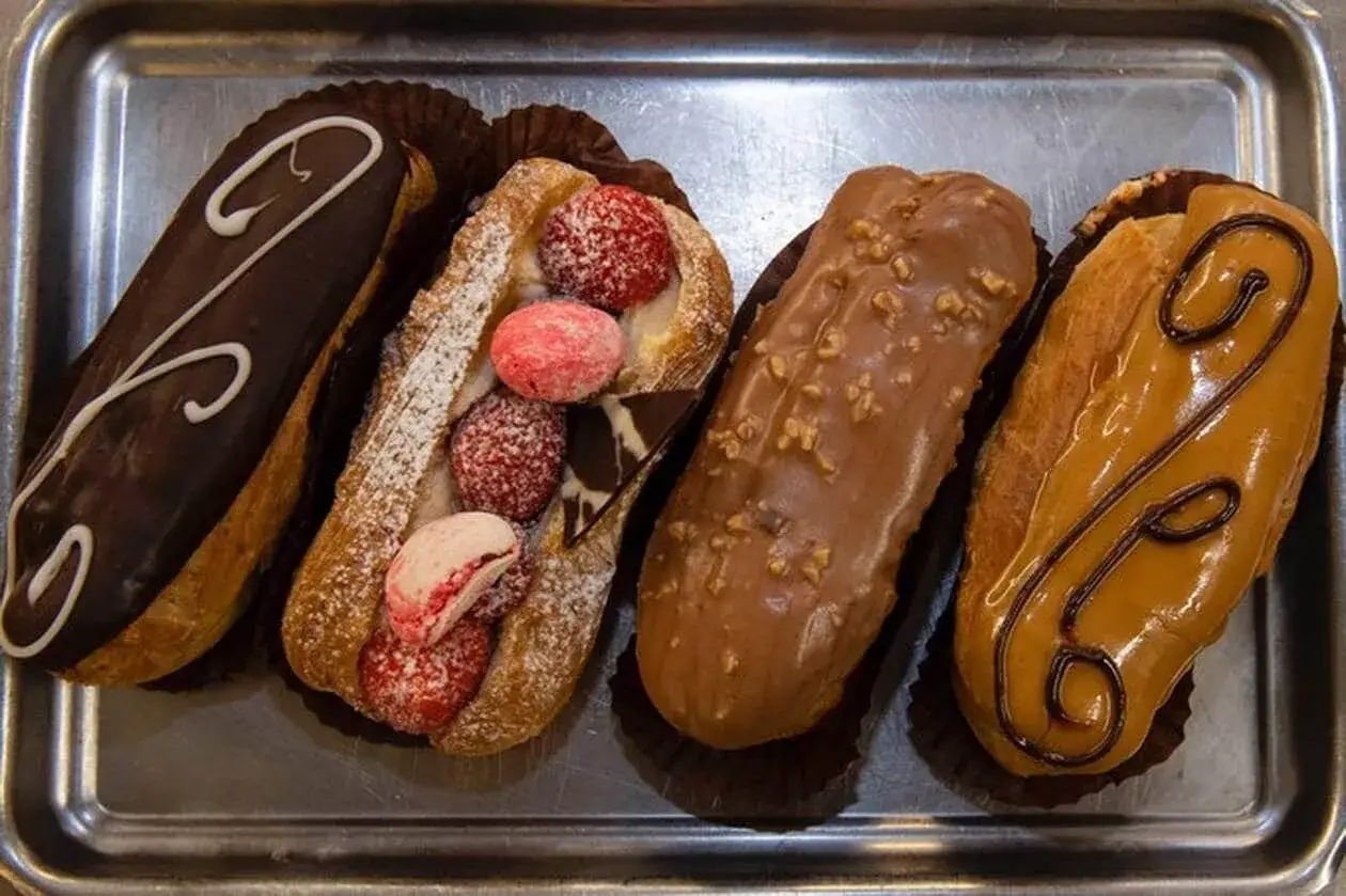 Quatre éclairs variés sur un plateau : un au chocolat noir, un garni de fruits rouges et macarons, un au chocolat praliné, et un à la crème caramel.