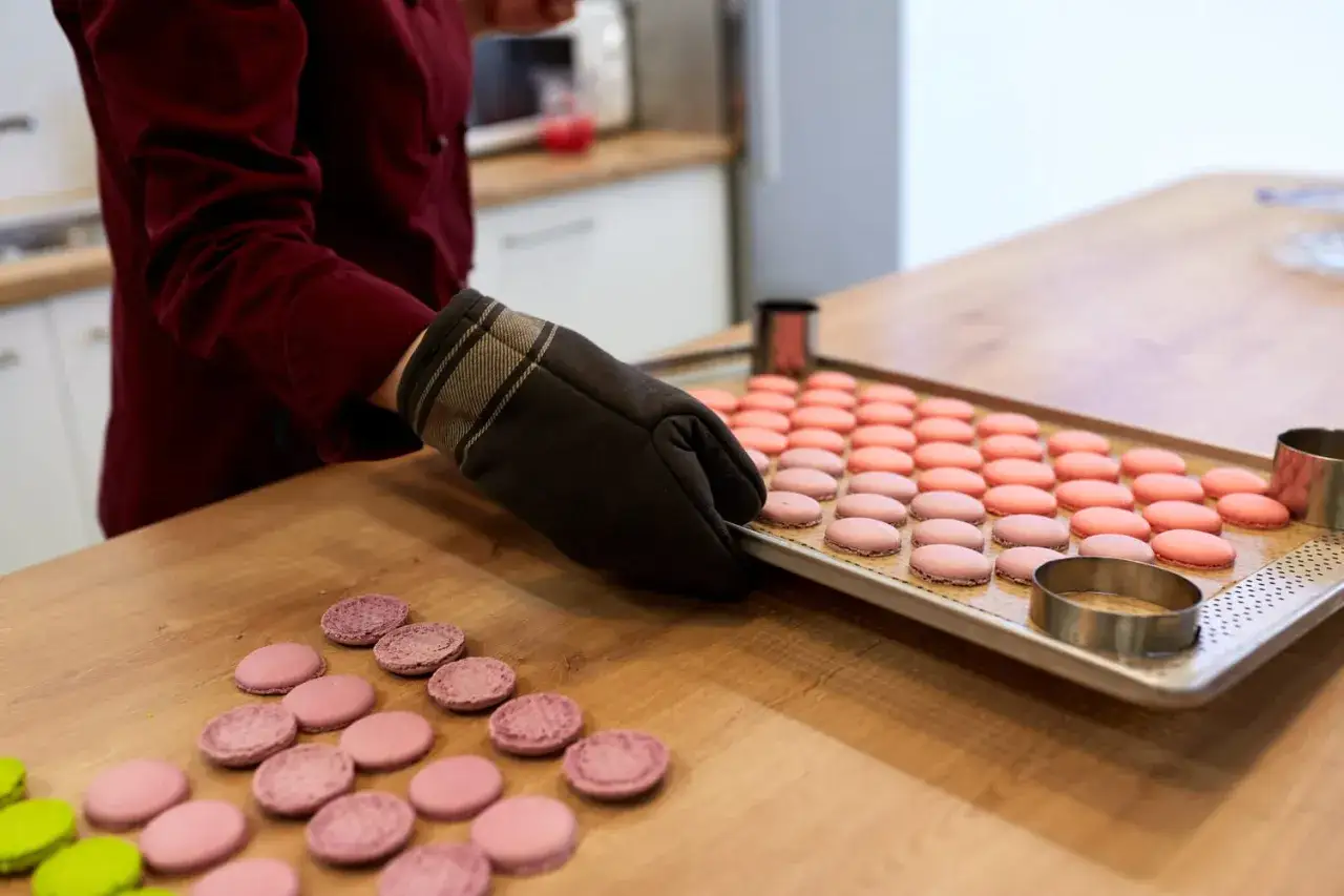 Pr&eacute;paration pour la cuisson macaron chaleur tournante : une main gant&eacute;e d&eacute;pose une plaque de macarons roses sur un plan de travail en bois.