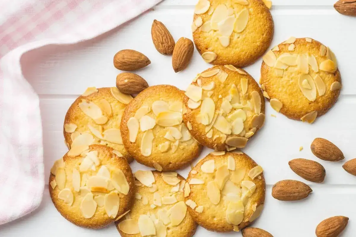 Des biscuits moelleux aux amandes, parsemés d'éclats d'amandes, sur un fond blanc avec quelques amandes entières et un tissu à carreaux roses.