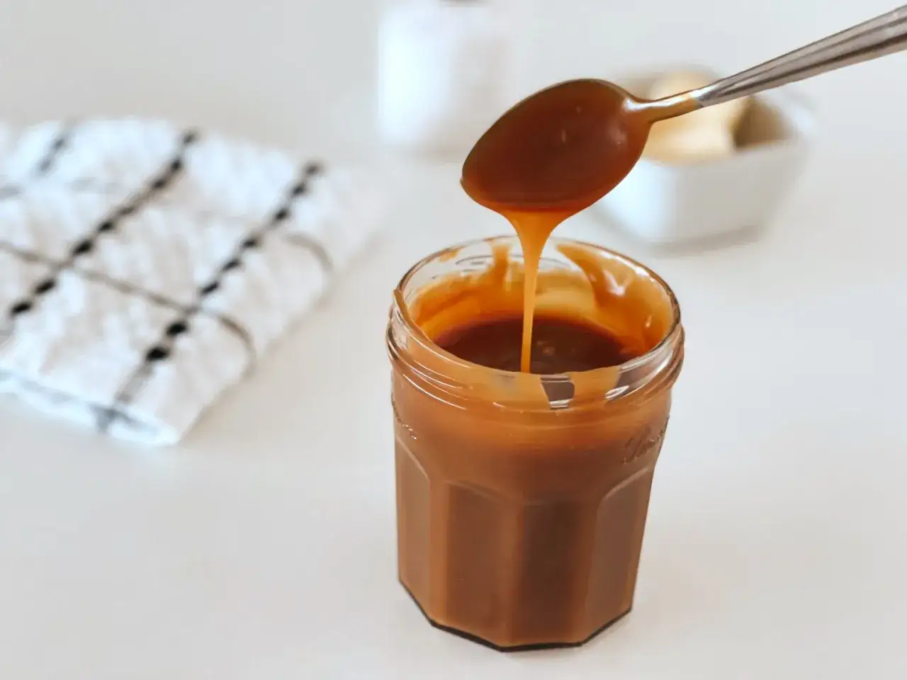 Caramel beurre sal&eacute; sans beurre demi sel coulant d'une cuill&egrave;re dans un pot en verre. Une serviette &agrave; carreaux est &agrave; gauche.