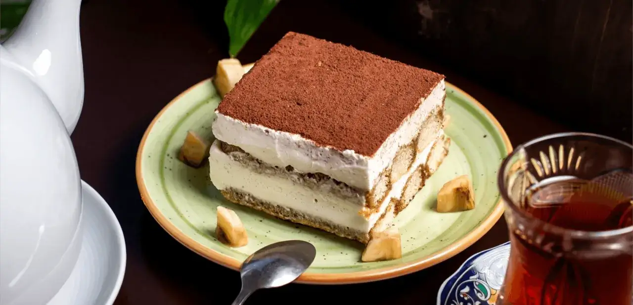 Une part de tiramisu onctueux, saupoudr&eacute;e de cacao, servie sur une assiette verte. Une tasse de th&eacute; ambr&eacute; et une th&eacute;i&egrave;re blanche compl&egrave;tent cette sc&egrave;ne digne d'une recette tiramisu chef &eacute;toil&eacute;.