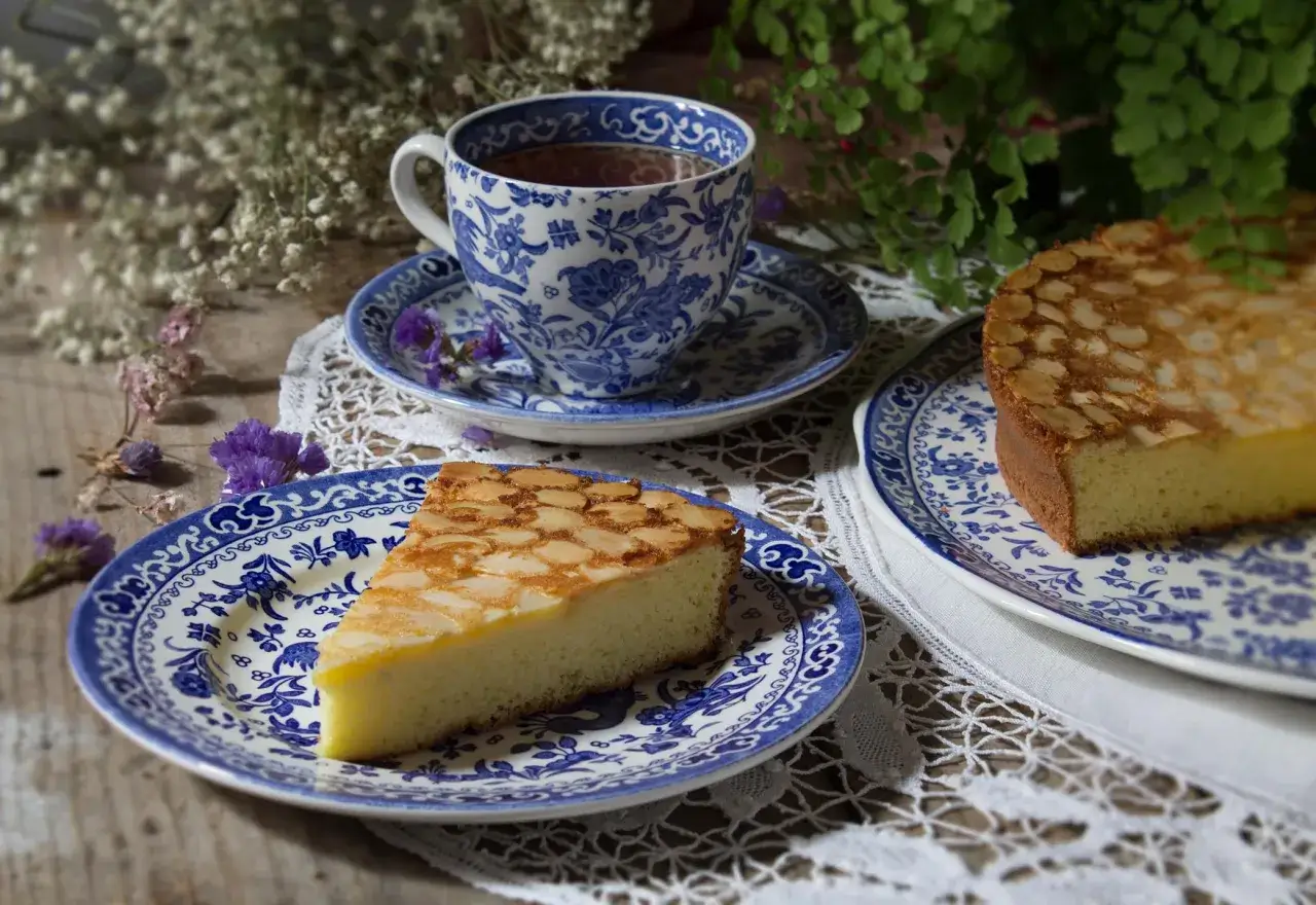 Une part de pain de gênes aux amandes dorées, servie sur une assiette bleue et blanche, avec une tasse de thé.