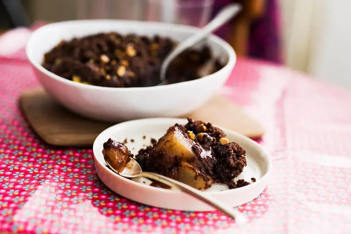 Un d&eacute;licieux crumble chocolat avec des morceaux de poire fondants, servi dans une assiette blanche sur une nappe rose &agrave; motifs.