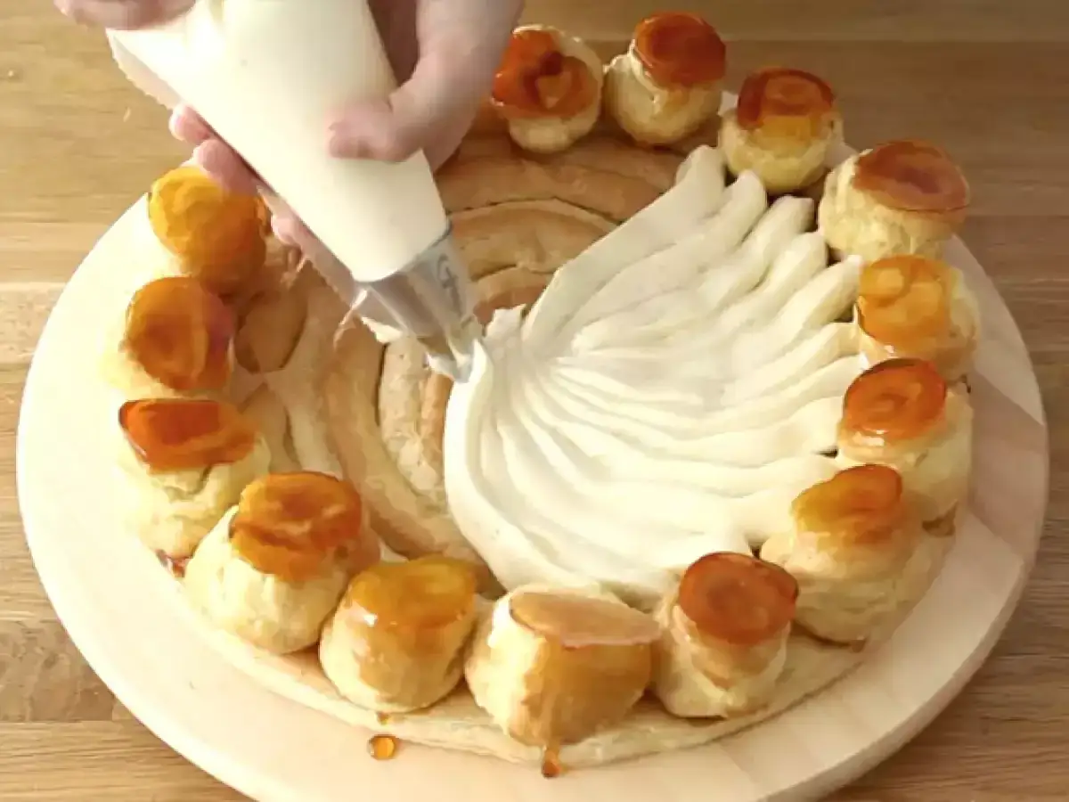 Une main garnit un gâteau rond de crème chiboust à l'aide d'une poche à douille. Des petits choux caramélisés ornent le pourtour.