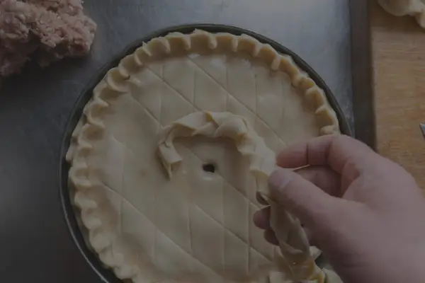 Main d'un pâtissier décorant une tarte avec une pâte brisée tressée. Un tas de farce est visible à gauche.
