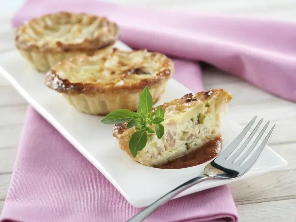 Une délicieuse petite quiche lorraine, garnie d'une touche de basilic frais, attend d'être dégustée sur une assiette blanche.