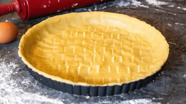 Un fond de tarte amande prêt à être garni, avec un rouleau à pâtisserie rouge et un œuf à proximité.