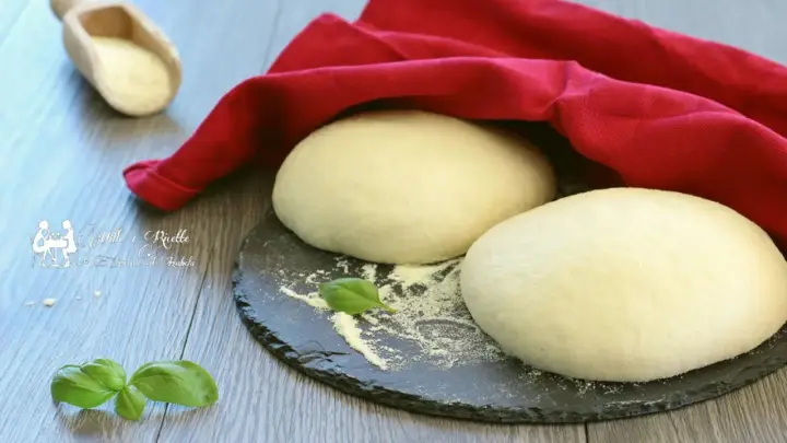 Deux boules de pâte à pizza professionnelle prêtes à être travaillées, sous un torchon rouge. Une feuille de basilic et une cuillère de semoule complètent la scène.