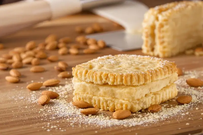 Trois biscuits joconde empilés, saupoudrés de sucre glace, entourés d'amandes sur une planche en bois.